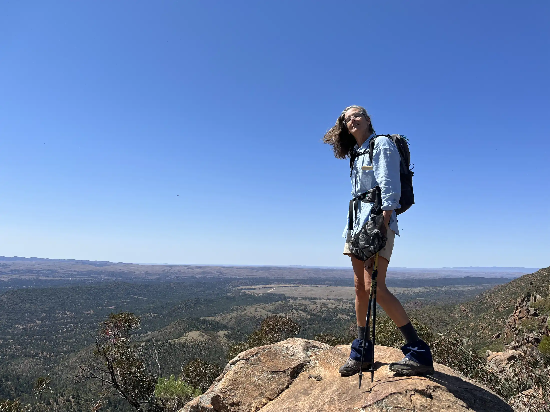 A woman wearing a backpack and holding hiking poles stands on a rocky mountaintop, looking out over a wide, sunlit landscape.